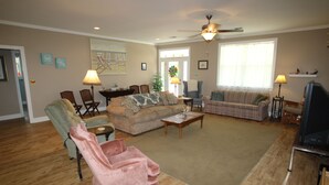 Ein geräumiges Wohnzimmer mit einem beigen Sofa, einem pinken Sessel und einem grünen Sessel. Es gibt einen Couchtisch, einen Fernseher und einen Deckenventilator.