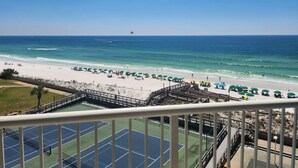 A beach view from a balcony with a tennis court in the foreground, a sandy beach with umbrellas, and the ocean in the background.