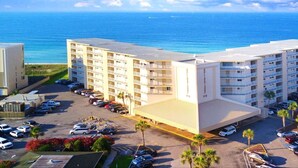 A multi-story hotel with a parking lot in front, located near a beach with the ocean in the background.