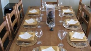 Una mesa de madera dispuesta para comer, con individuales, platos, cubiertos y copas de vino.