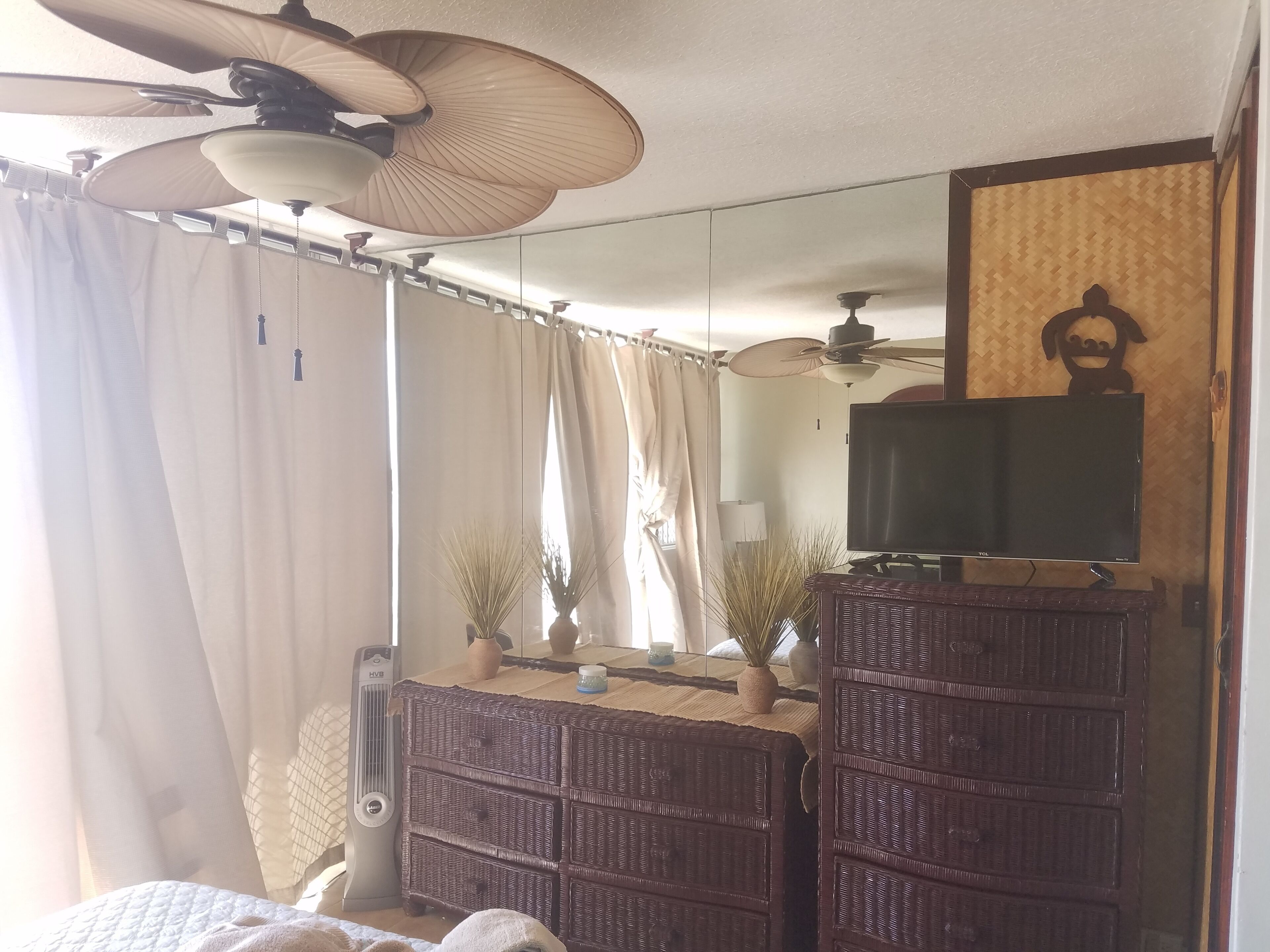 A hotel room with a bed, a TV on a stand, a ceiling fan, a mirror, and a window with curtains.
