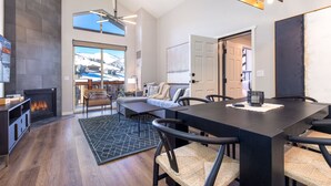 A modern living room with a fireplace, a dining area with a black table and chairs, and a large window with a view of a snowy landscape. - Ski In/Out 2BD Top floor W/Amazing Views (Park City)