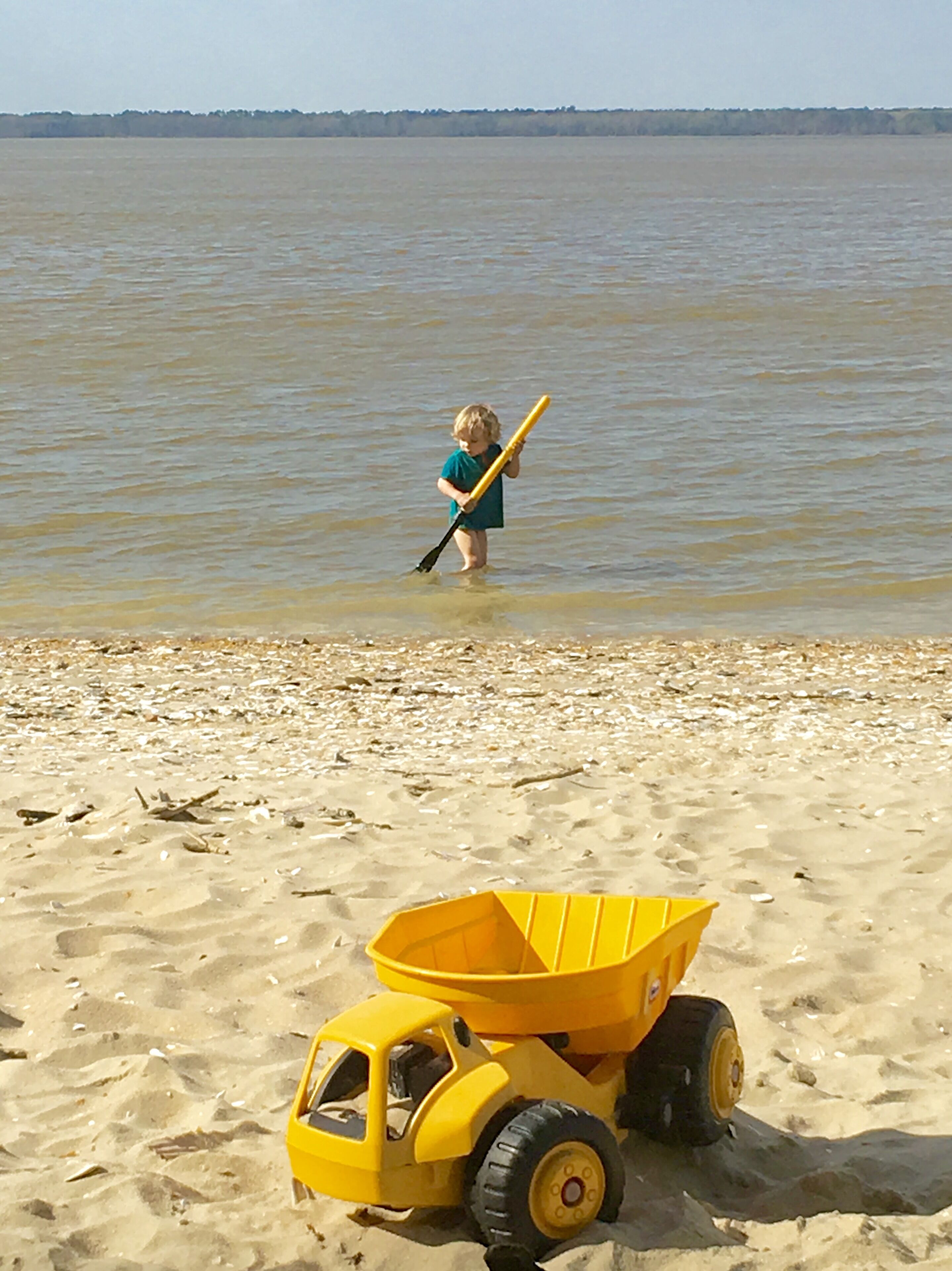 A child playing near a beach with a toy dump truck and a toy paddle in the water.