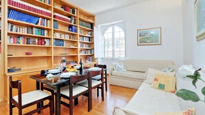 A living room with a wooden bookshelf, a dining table with chairs, a sofa, and a window with a painting on the wall. - Rome Colosseum Vacation Rental - Spacious Apartment Ancient Rome 3BR/2B Wi-Fi (Roma)