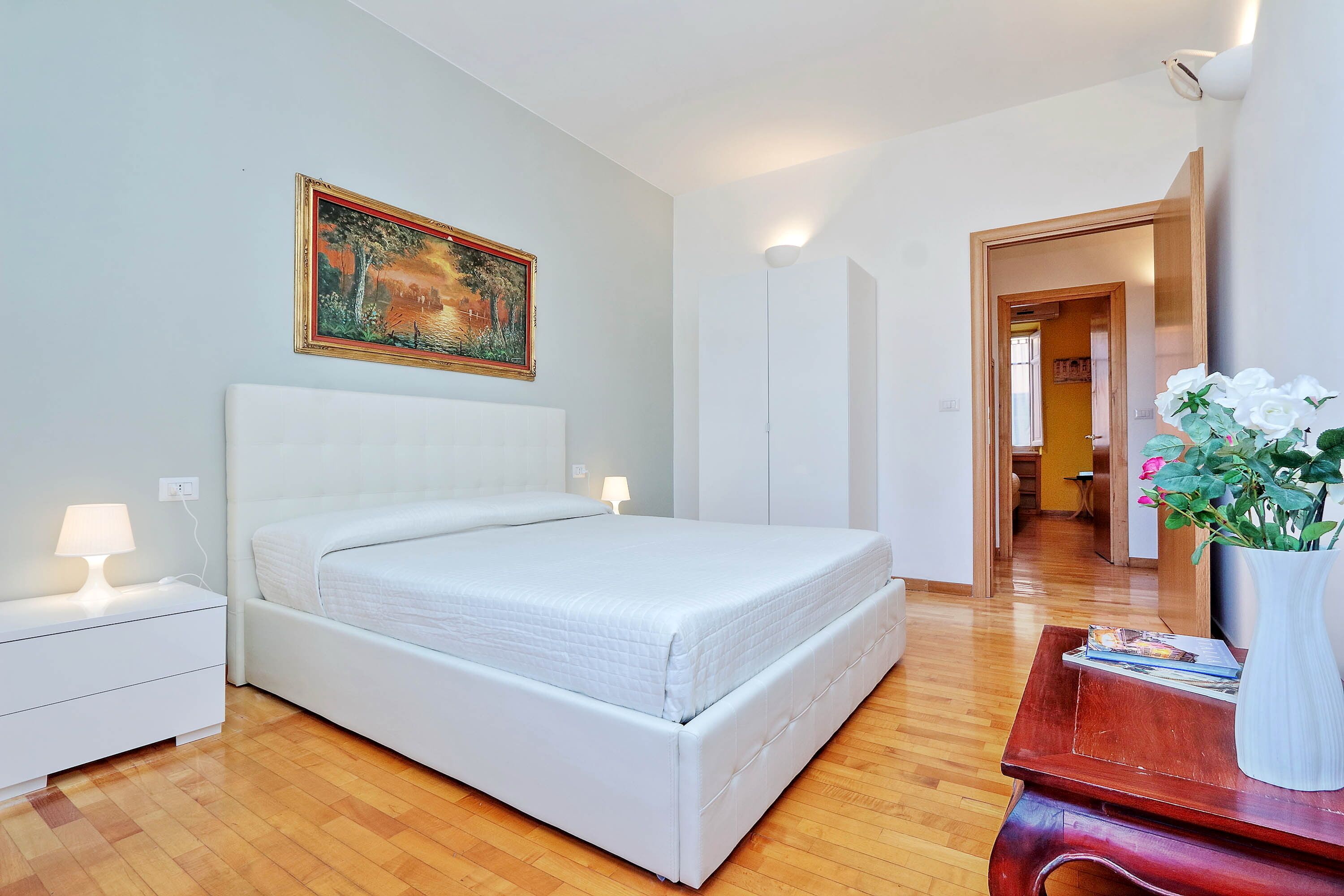 A bedroom with a bed, bedside tables, a nightstand, a painting, a wooden floor, and a door leading to another room.
