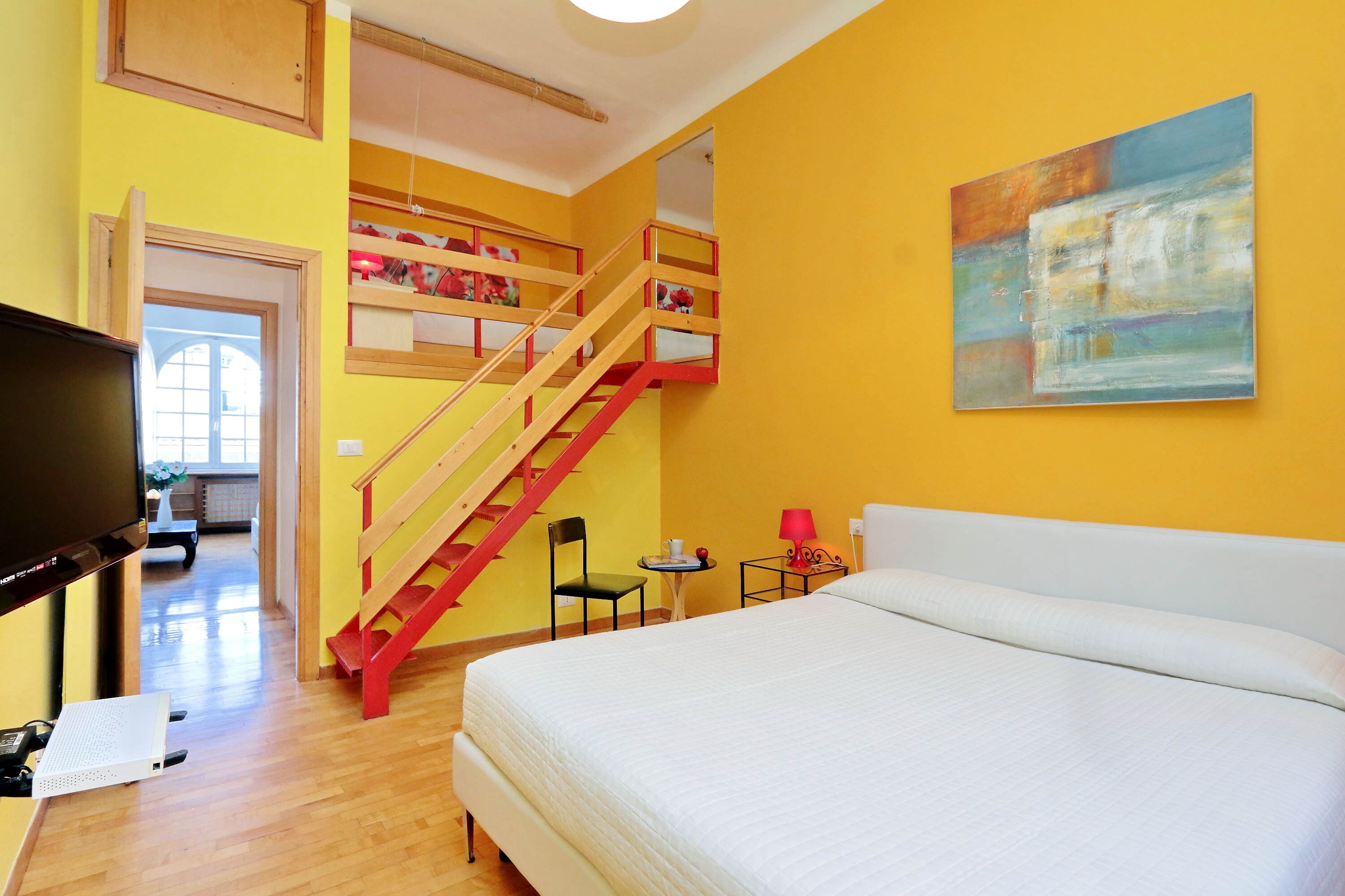Habitación de hotel con cama, televisor, escritorio, silla, mesita, lámpara y un cuadro en la pared.