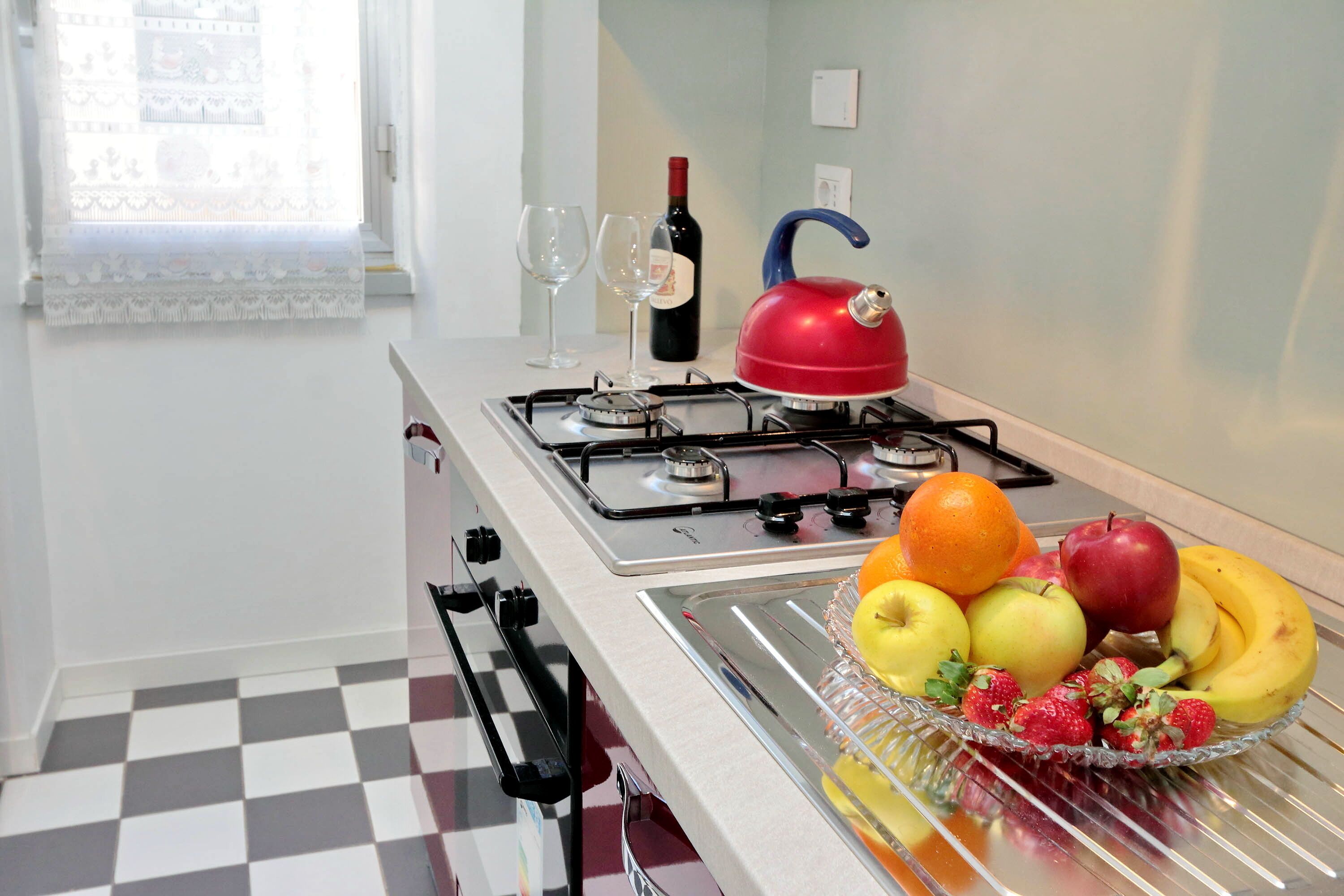 Een keuken met een gasfornuis, een rode waterkoker en een fruitschaal met appels, bananen en aardbeien.