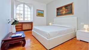A bedroom with a large bed, a window with curtains, a bedside table with a lamp, and a painting on the wall. - Rome Colosseum Vacation Rental - Spacious Apartment Ancient Rome 3BR/2B Wi-Fi (Roma)