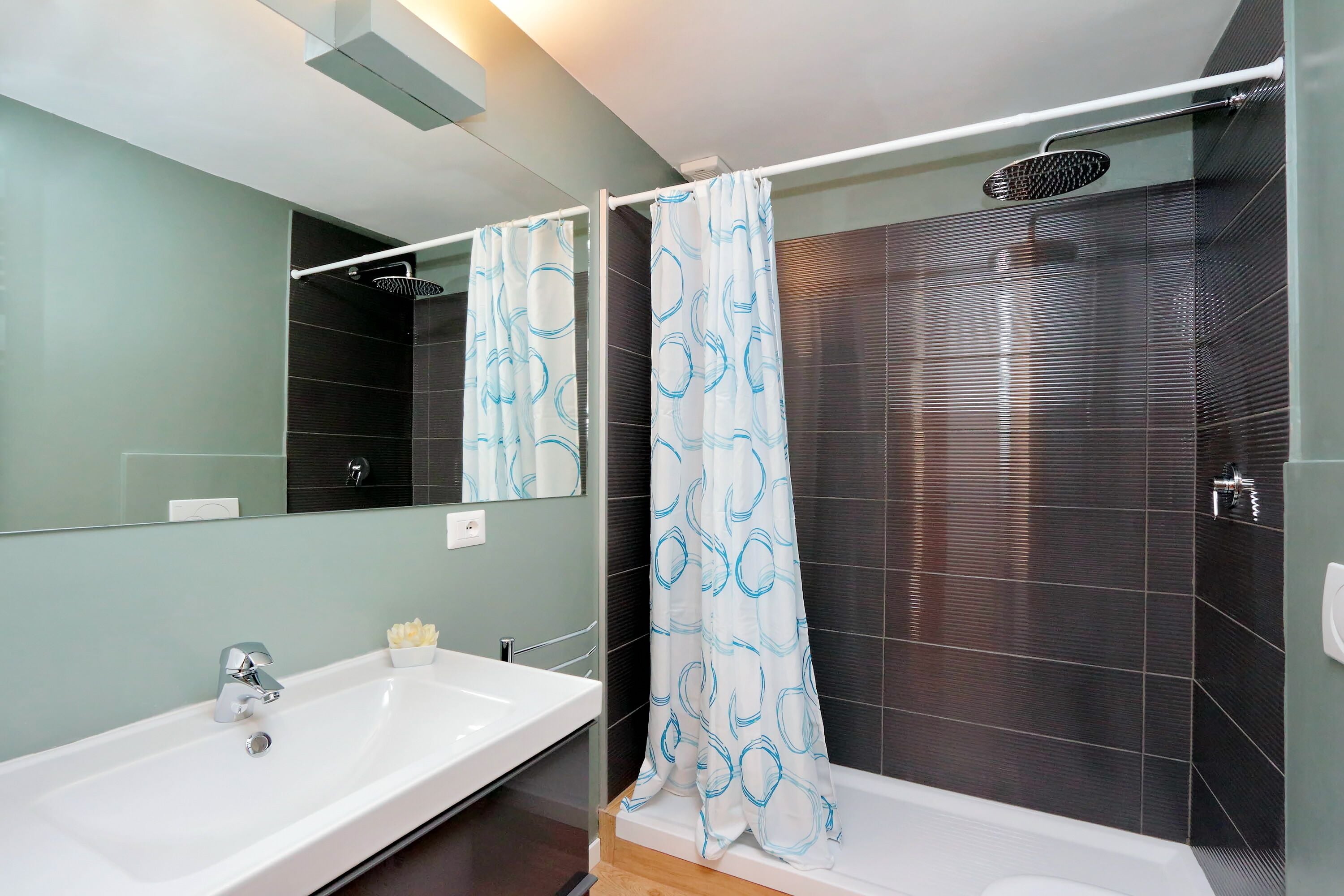 Een moderne badkamer met een wit wastafel, een spiegel, een douche met gordijn en donkere betegelde wanden.