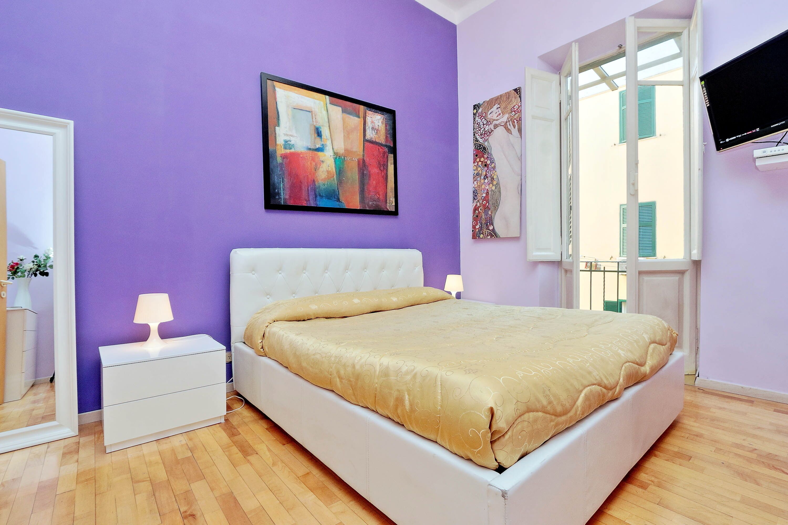 A bedroom with a white bed, a nightstand, a lamp, a painting, and a window with a view of buildings.