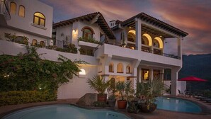 Una casa de dos pisos con piscina, zona de estar exterior y montañas al fondo.
