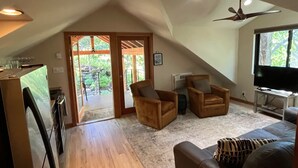 A spacious living area with a high ceiling, a ceiling fan, a flat-screen TV, and a large window providing a view of the outdoors.