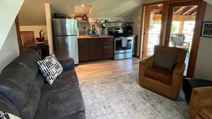 A compact kitchen with modern appliances, a dining area with a sofa and armchair, and a view through a glass door to an outdoor patio.