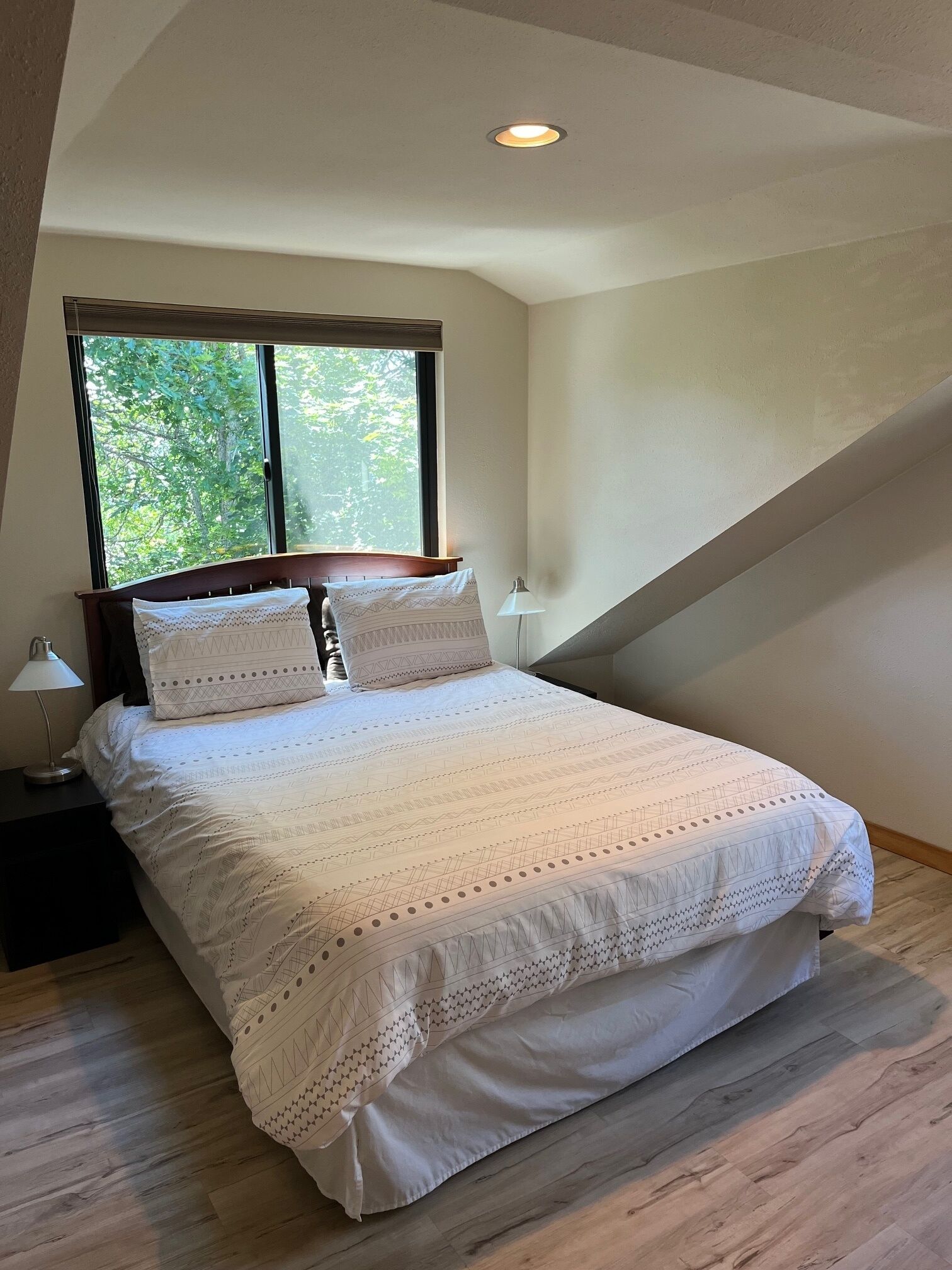 Une chambre à coucher avec un grand lit, une table de chevet, une lampe et une fenêtre donnant sur des arbres.