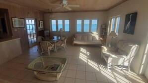 A spacious living area with a wooden ceiling, a ceiling fan, a dining table with chairs, a sofa, and a large window overlooking the sea. - Centrally located - Gorgeous view of Elizabeth Harbour  (George Town)