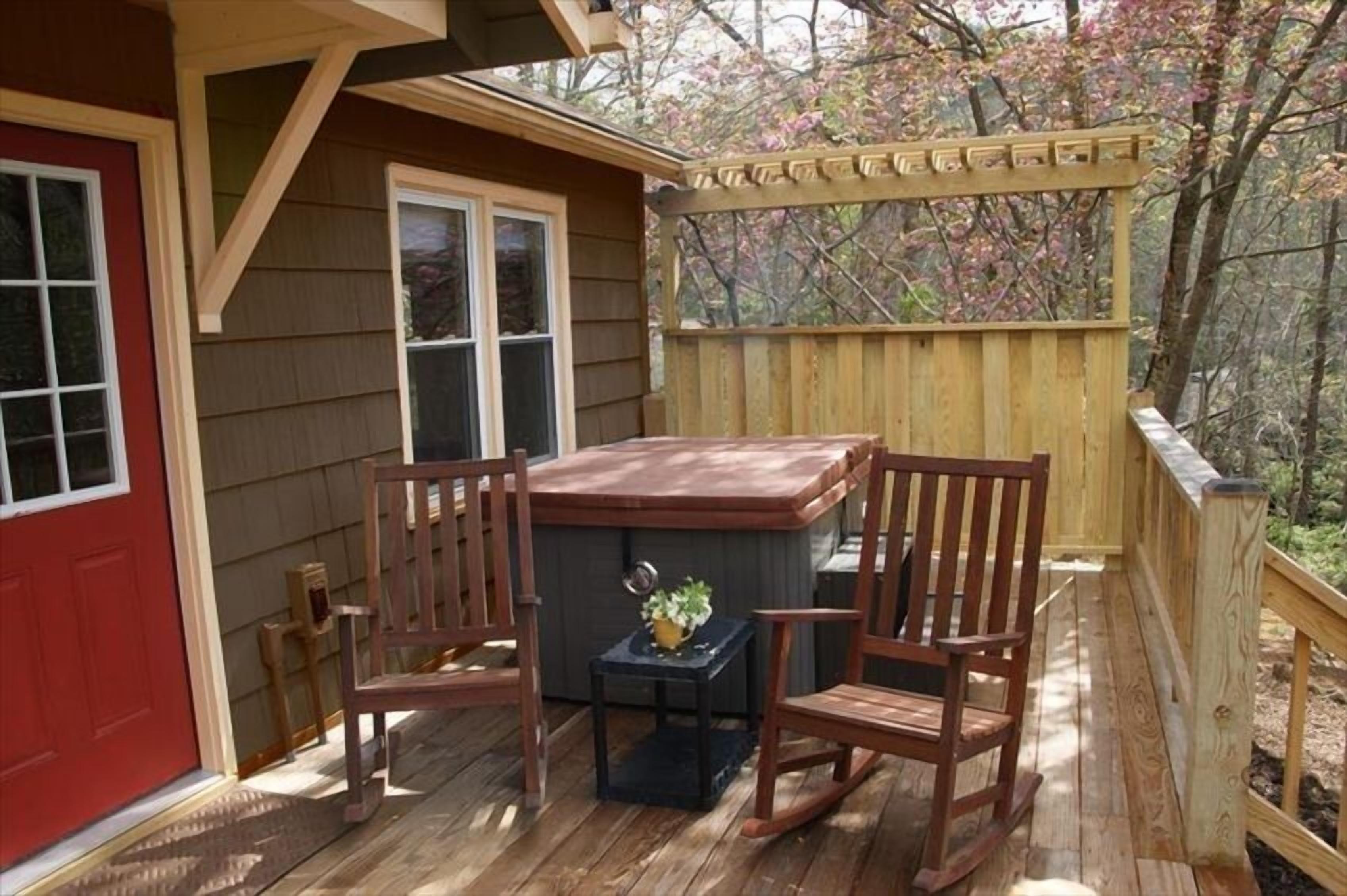 A wooden deck with a hot tub, two rocking chairs, and a small table with a plant.