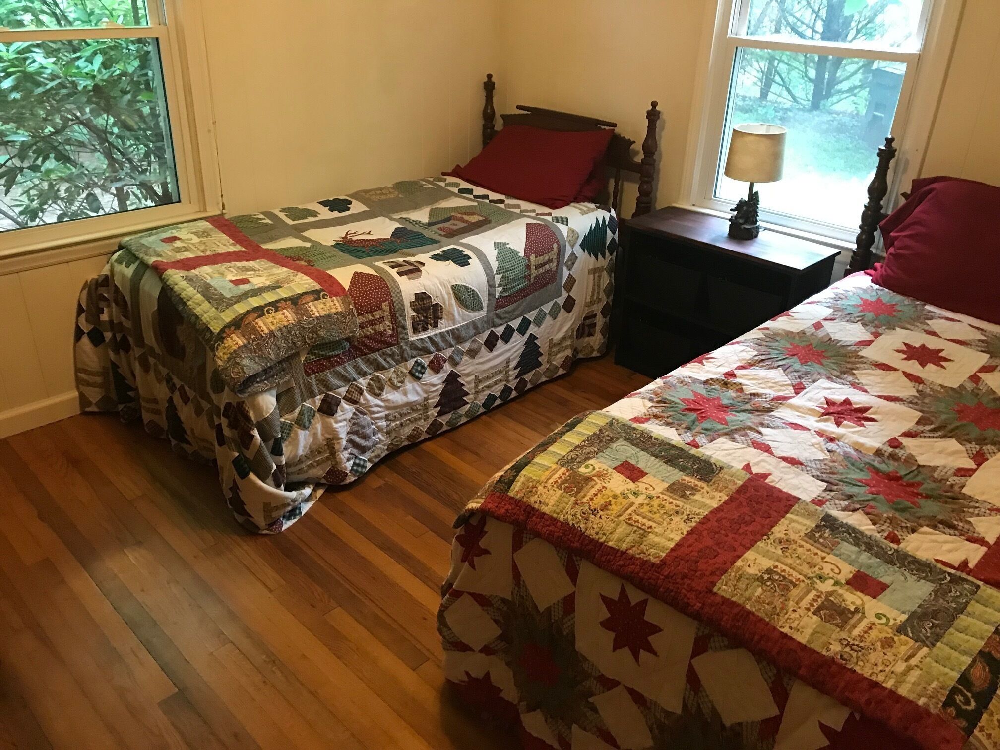 A room with two beds covered in quilts, wooden flooring, and a window with a view of trees.