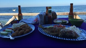 Ein Tisch ist reichhaltig mit Speisen gedeckt, darunter Reis, Fisch und Gemüse, sowie zwei Bierflaschen. Im Hintergrund ist das Meer zu sehen.