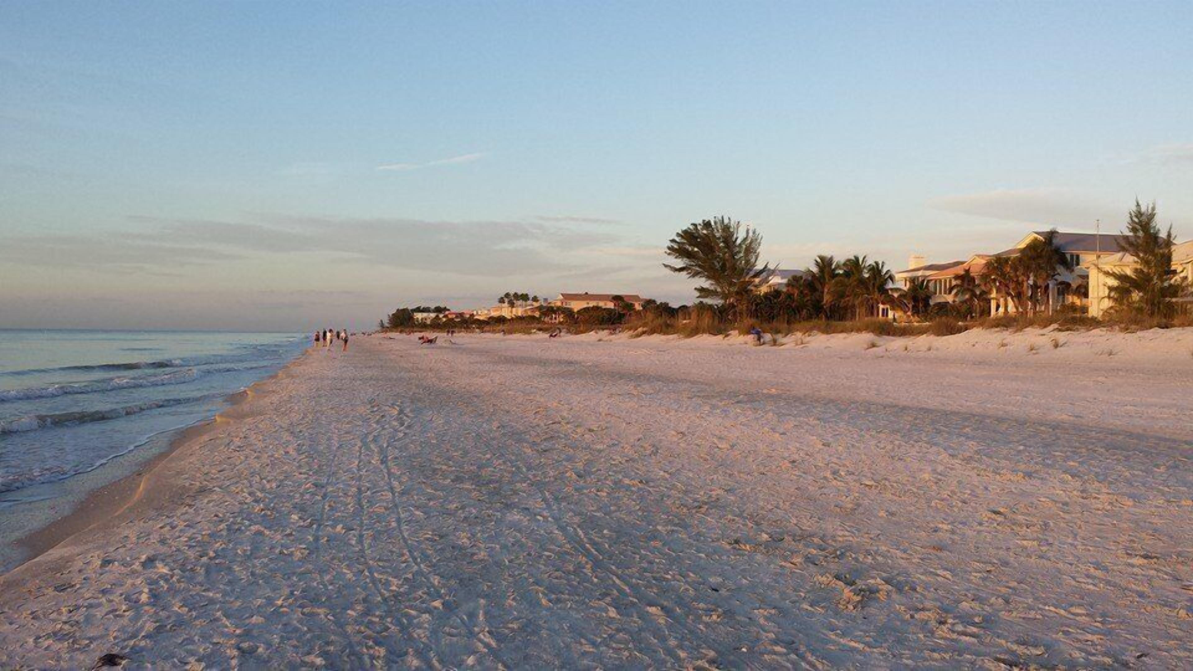 Una playa amplia y arenosa con algunas personas caminando, un mar tranquilo y casas al fondo.