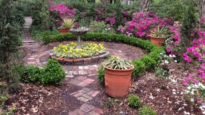 A garden with a brick pathway, a central fountain, and various potted plants and flowers.