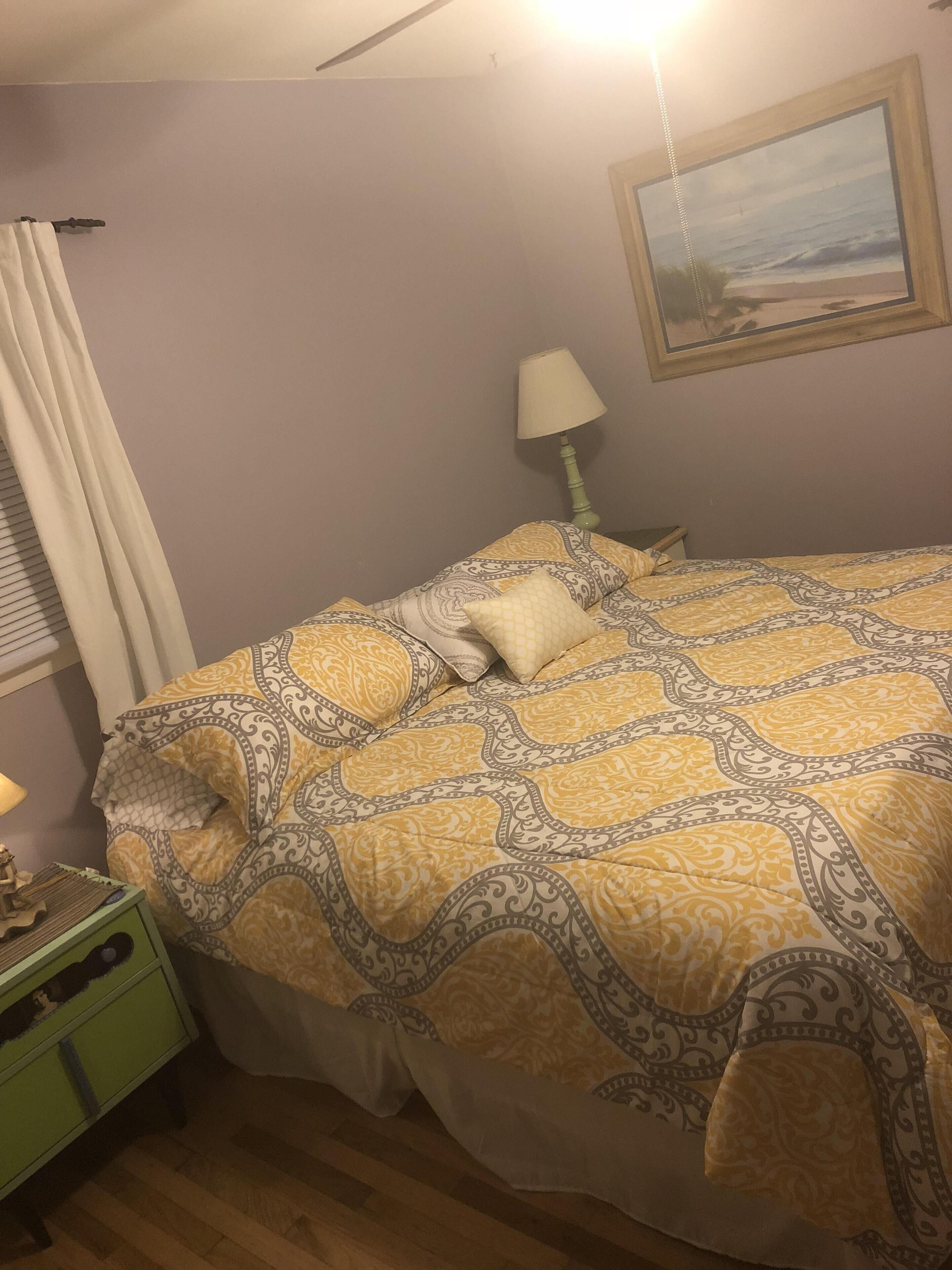 Une chambre à coucher avec un lit recouvert d’une literie à motifs, une table de chevet avec une lampe, une commode verte et un tableau accroché au mur.