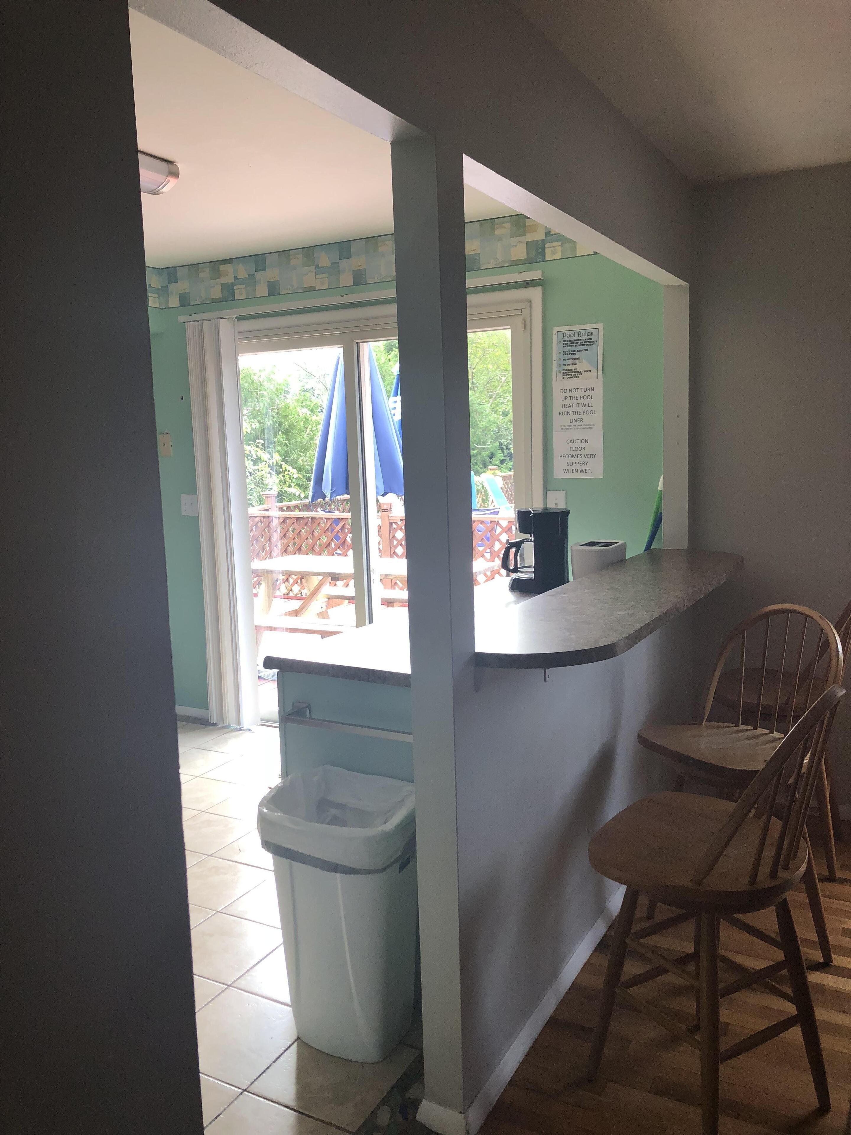 A kitchen area with a counter, two wooden chairs, a trash bin, and a view of an outdoor deck with umbrellas.