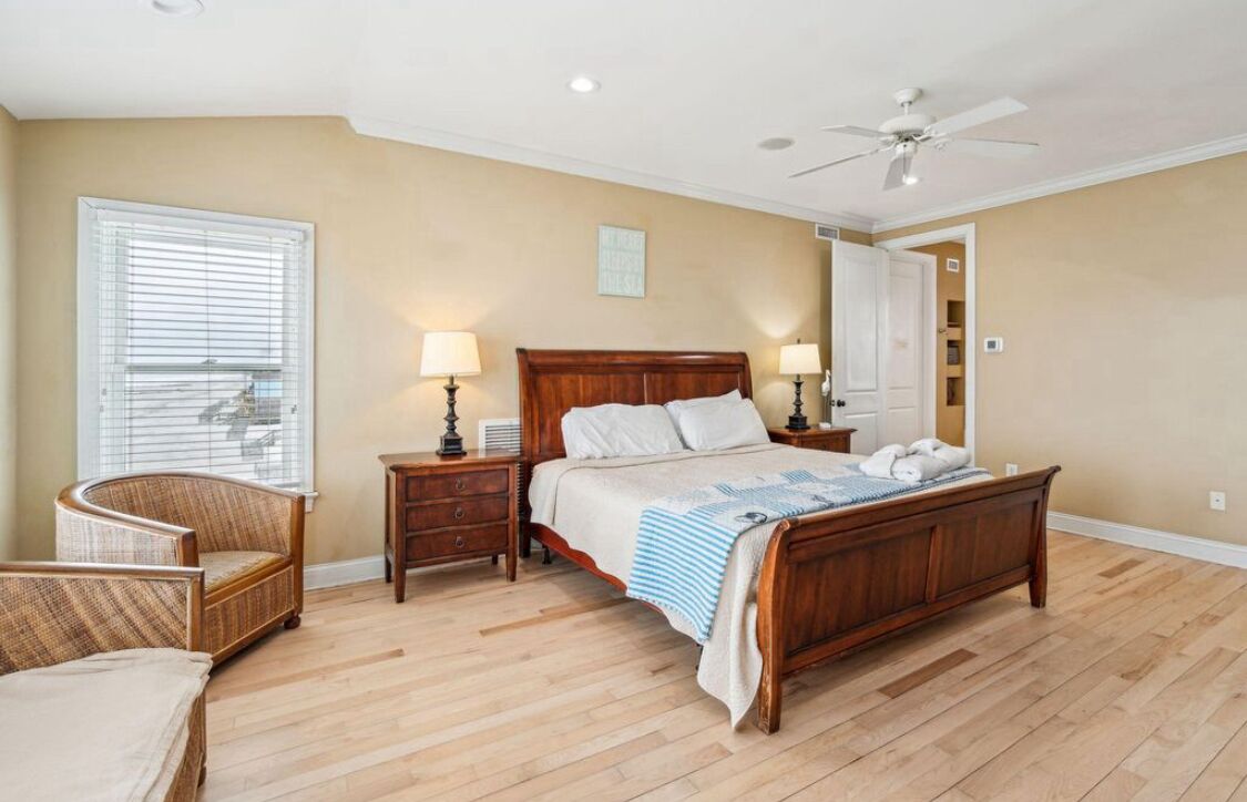 A bedroom with a wooden bed, two bedside tables with lamps, a chair, a window with blinds, and a ceiling fan.