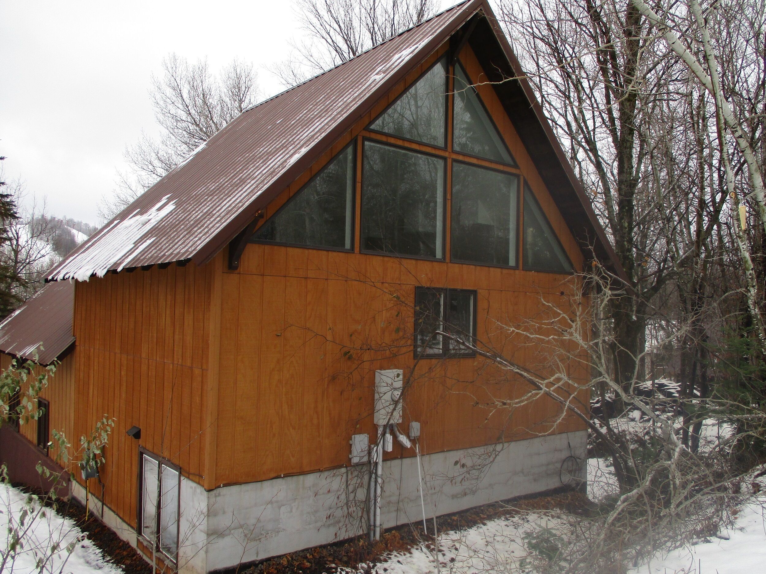 A wooden cabin with a sloped roof and large windows, surrounded by trees and snow.