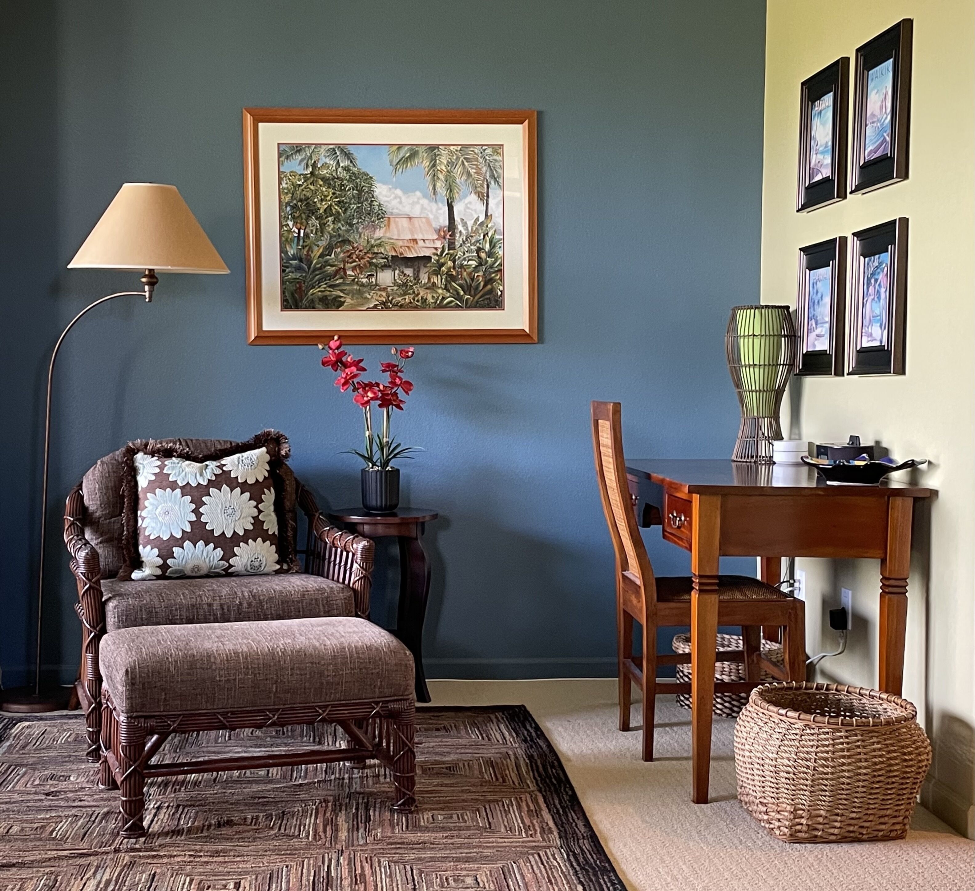 A room with a dark blue wall, a framed landscape painting, a wooden chair with a cushion, a small table with a vase of flowers, and a lamp.