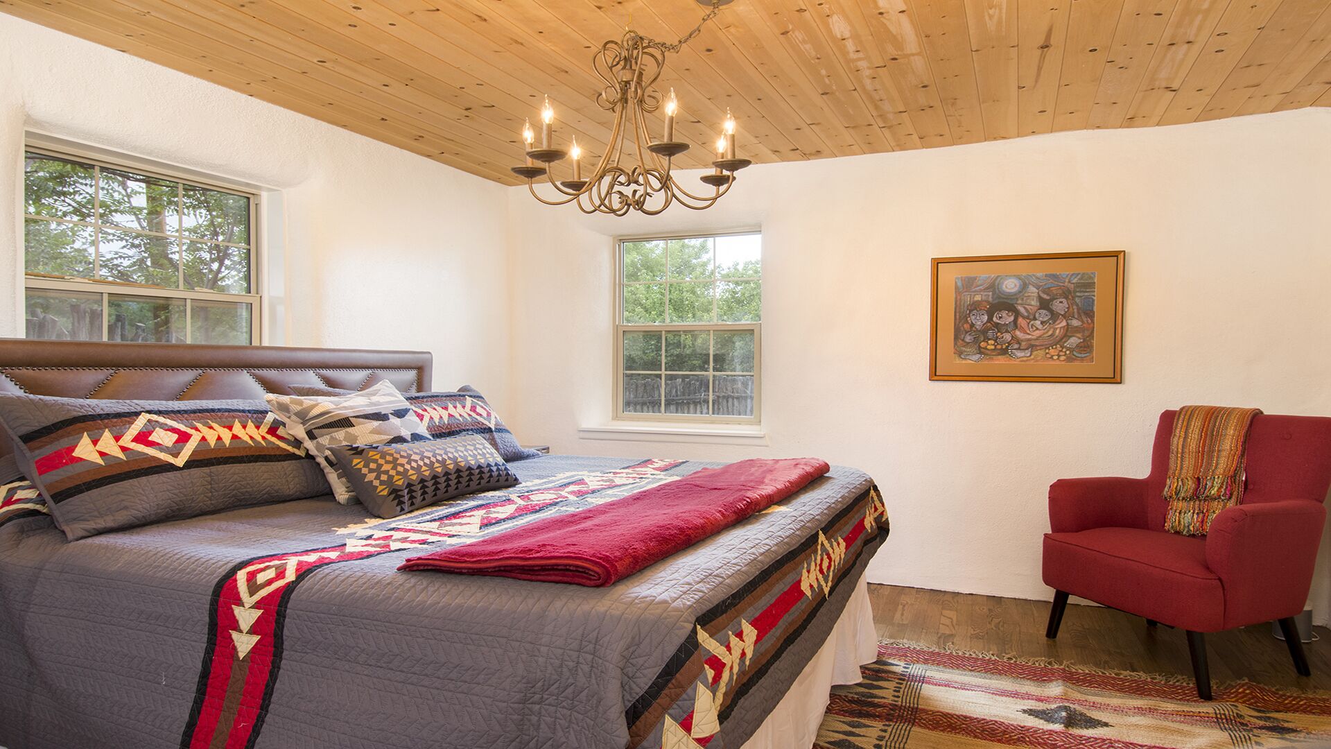 Un dormitorio con una cama grande, un sillón rojo, una araña y una ventana con vista a árboles.