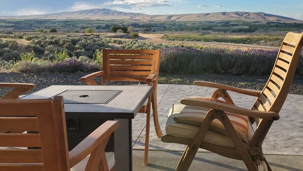 Un patio con muebles de madera y una mesa, con vistas a un paisaje de colinas y flores moradas.