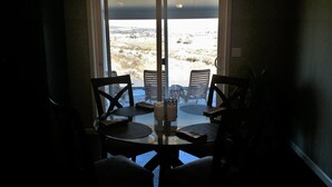 Comedor con mesa de vidrio y cuatro sillas de madera, que da a un patio exterior con sillas y vistas a un paisaje rural.