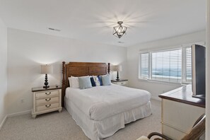 A bedroom with a wooden headboard, two bedside tables with lamps, a large bed with white bedding, a window with blinds, and a television on a stand. - Oceanfront Views, Community Pool, Great Resort Location! (Isle Of Palms)