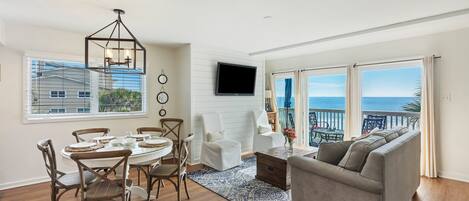 A spacious living room with a large window overlooking the ocean, a dining area with a round table, and a gray sofa.