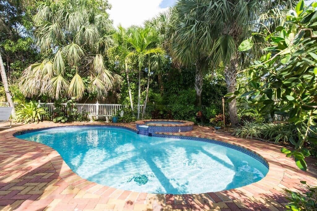 A swimming pool with a tiled edge, surrounded by lush greenery and a brick pathway.