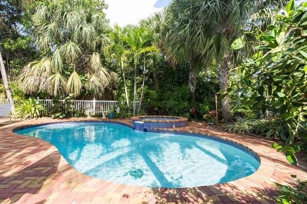 A swimming pool with a tiled edge, surrounded by lush greenery and a brick pathway.