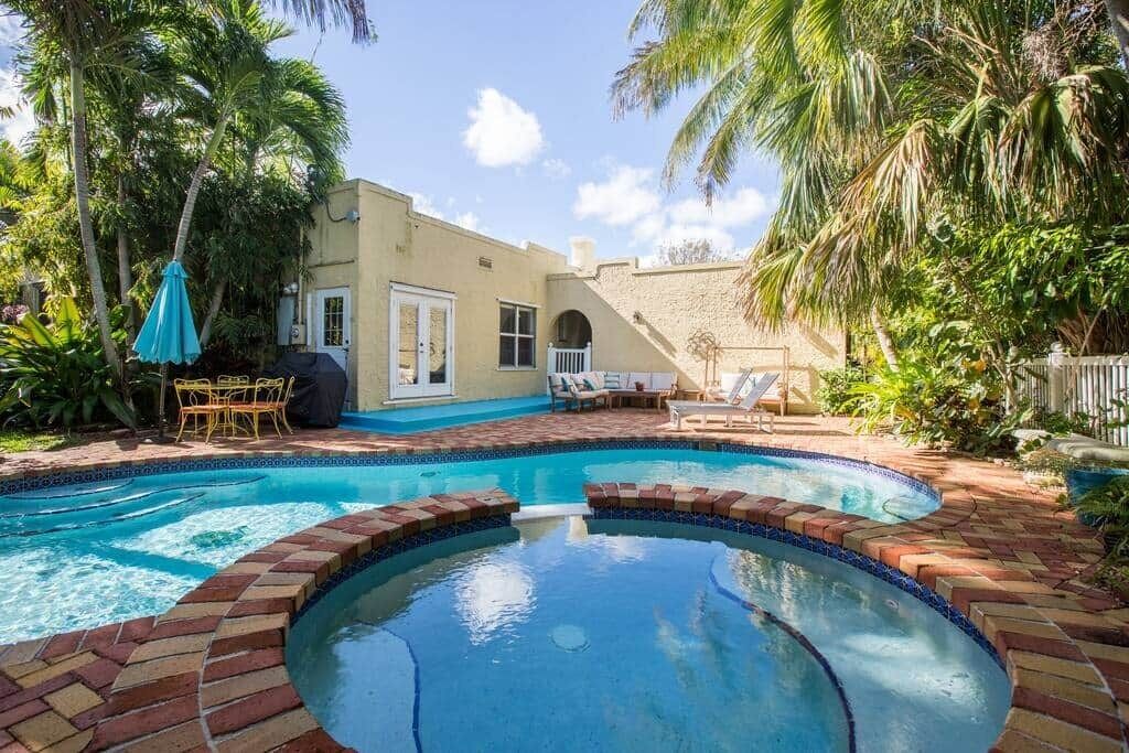 Una piscina con borde de ladrillo, rodeada de palmeras y un patio con mobiliario exterior.