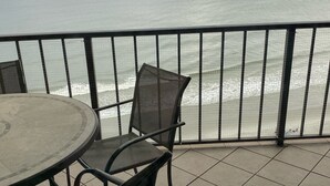 Een balkon met een ronde tafel en twee stoelen, met uitzicht op een strand met golven en een bewolkte lucht.
