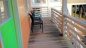 A covered wooden deck with a bench and a chair, a white railing, and a view of a building with windows. - One Street Over - Cottage by the Sea! (Carolina Beach)