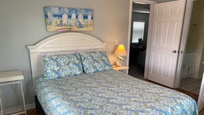A bedroom with a bed, a nightstand, a lamp, a painting, and a door leading to another room. - One Street Over - Cottage by the Sea! (Carolina Beach)