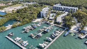 An aerial view of a marina with boats docked, a large building with multiple floors, and surrounding greenery.