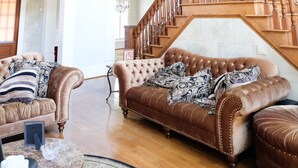 Un vaste salon doté d’un haut plafond, d’un lustre, d’un escalier avec des rampes en bois, et de deux canapés en cuir marron agrémentés de coussins décoratifs à motifs.