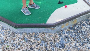 Um campo de minigolfe com superfície de jogo verde, uma bola vermelha e um jogador com camiseta branca e bermuda azul.
