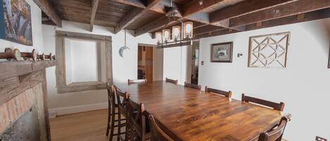A spacious dining area with a long wooden table and chairs, a rustic ceiling, and a brick wall.