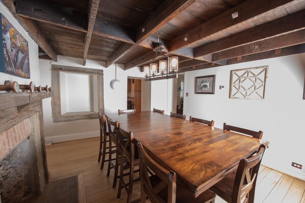 A spacious dining area with a long wooden table and chairs, a rustic ceiling, and a brick wall.