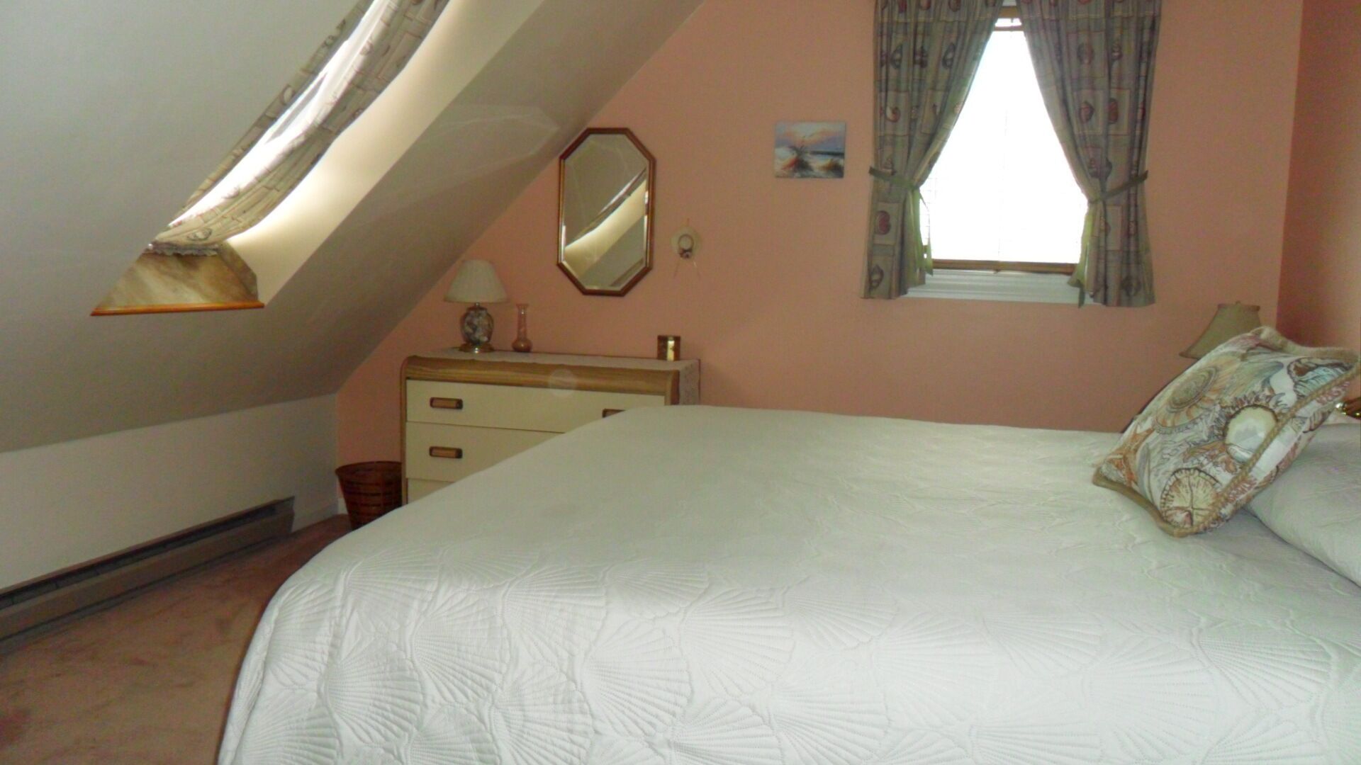 Une chambre avec un plafond en pente, un lit agrémenté d’un coussin à motifs, une commode, un miroir et une fenêtre avec des rideaux.