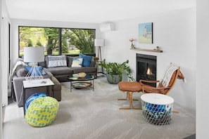Una sala de estar moderna con un sofá, un sillón y una mesa de centro, una chimenea y ventanas amplias con vistas a árboles.