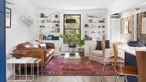 A compact living space with a brown leather sofa, a dining area with wooden chairs, a bookshelf filled with books and decorative items, and a large window with a view of greenery. - River's Edge (Leland)