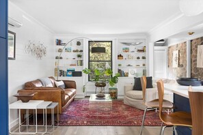 A compact living space with a brown leather sofa, a dining area with wooden chairs, a bookshelf filled with books and decorative items, and a large window with a view of greenery. - River's Edge (Leland)