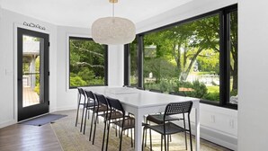 A modern dining area with a white table and black chairs, large windows with a view of greenery, and a textured pendant light. - River's Edge (Leland)
